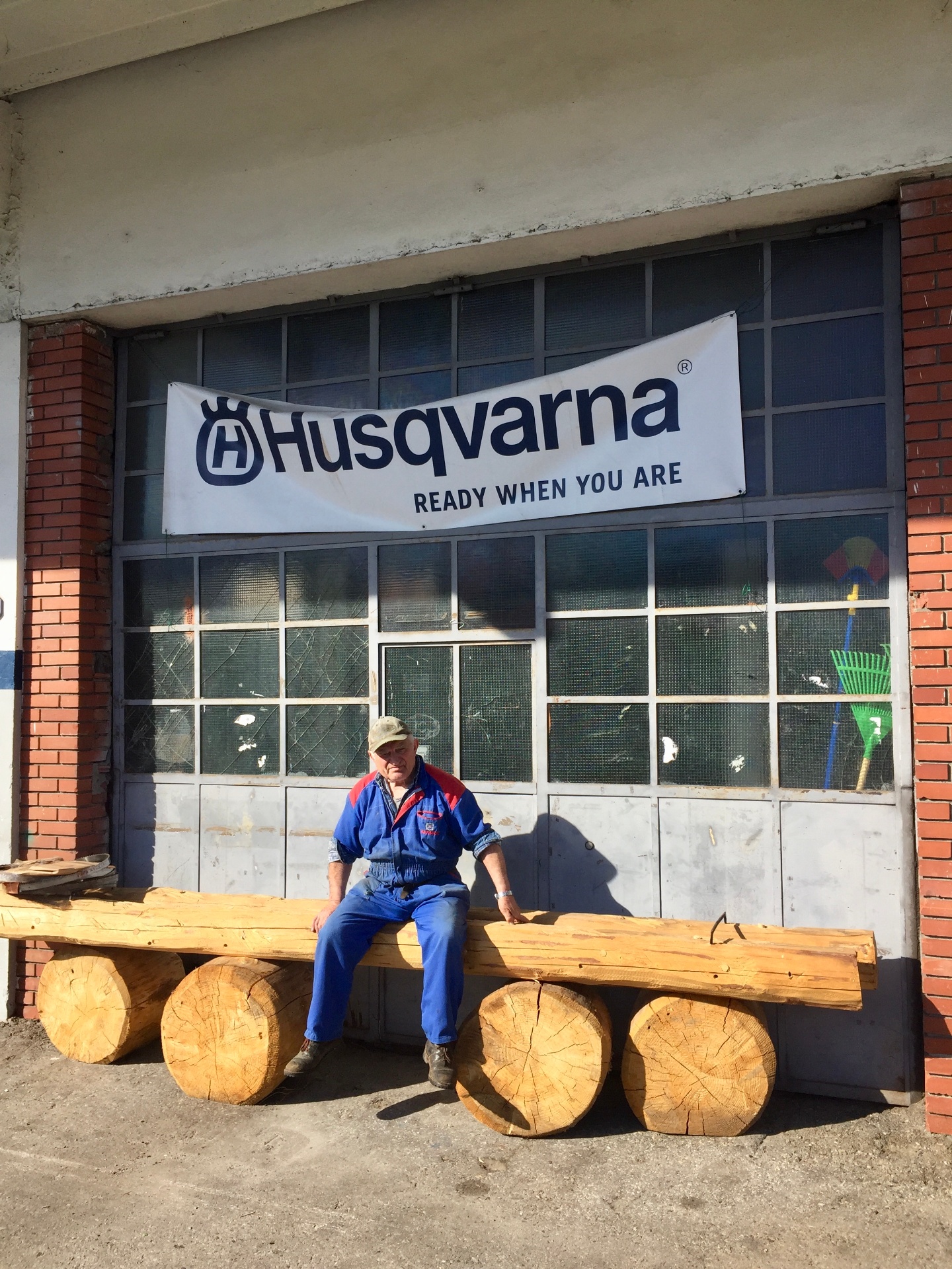 Storico titolare Papa Nicolini seduto su una panca in tronchi davanti all'officina, sotto lo striscione Husqvarna Ready When You Are